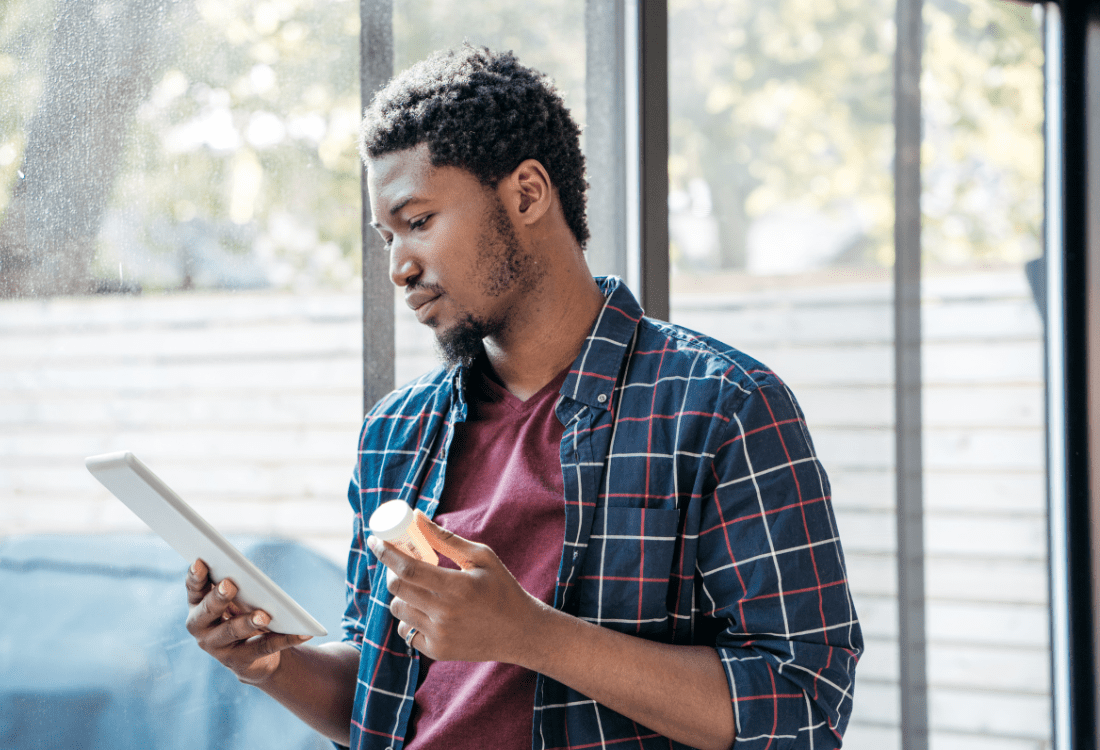 A man reads information on a tablet while holding a bottle of medication in his hand, grateful that he has the option for discrete management of health issues through the use of a trusted online pharmacy.