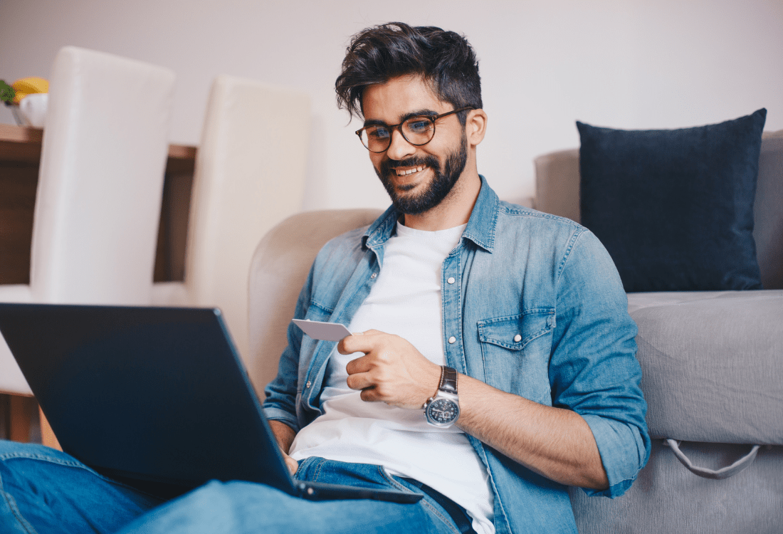 A cheerful man sits comfortably on his sofa whilst browsing his laptop and researching alternative ED medications to Viagra with privacy and ease. 