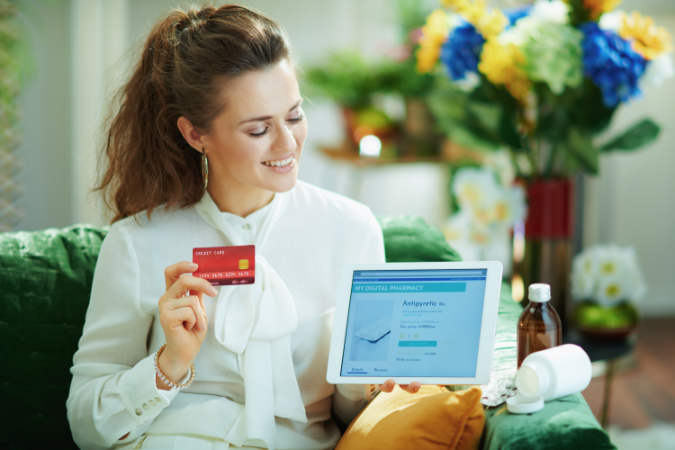 A woman sitting comfortably on a green sofa and smiling as she completes her order for medication via an online pharmacy, emphasising the ease and convenience in managing everyday health issues through online services. 